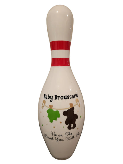 A white baby shower bowling pin with custom printed text reading 'Baby Broussard' and 'He or She Loved You Will Be', decorated with a green shirt, brown teddy bear, and clothesline details.
